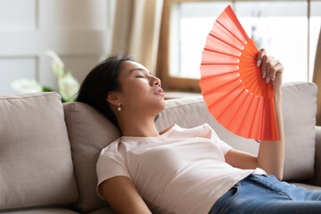 Overheated exhausted Asian woman waving orange paper fan, sitting on couch at home, tired girl...