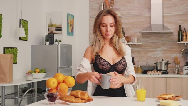 Attractive blonde lady in lingerie holding a cup of coffee drinking during breakfast enjoying the morning. Young sexy woman with tattoos in seductive underwear relaxing in the kitchen smiling.