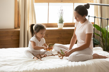 Caring young Asian mother and little daughter playing toy dinosaurs, sitting on cozy bed together,...