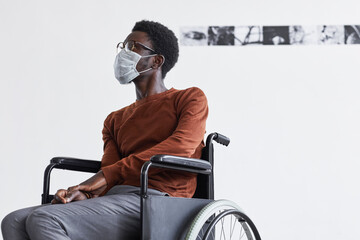 Minimal portrait of African-American man using wheelchair and wearing mask while looking at paintings in modern art gallery, copy space