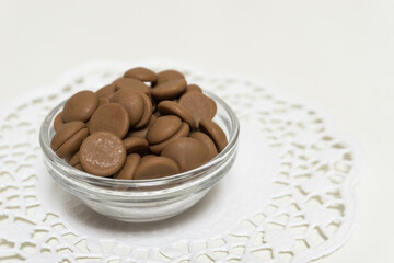 Many brown chocolate pellets in a glass bowl on white background