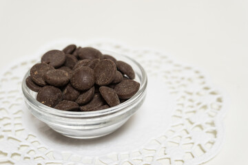 Many dark brown chocolate pellets in a glass bowl on white background