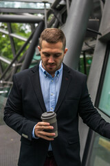 Photo of a young and attractive business man enjoying his coffee break outdoors. He is wearing smart clothes and is drinking in a reusable cup	