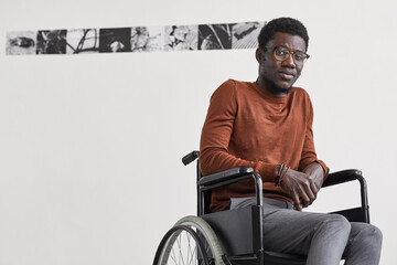 Minimal portrait of young African-American man using wheelchair and looking at camera while posing against white wall in modern art gallery, copy space