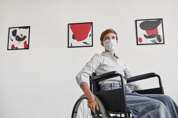 Wide angle portrait of young woman using wheelchair and wearing mask while looking at paintings in modern art gallery