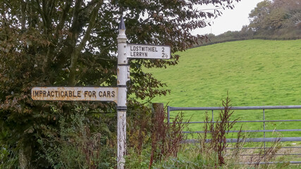 Old, public cast iron signpost, with fingerpost direction showing as Impracticable for cars. In rural setting with metal gate and hillside in background. Landscape image with space for text. Cornwall. - 384815641
