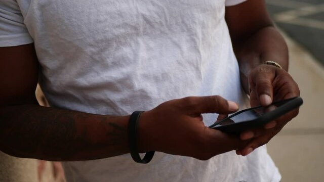 African American man on his phone texting and searching the internet in a white shirt and gold chain around his neck, next to brick and mortar building as camera pans up. Shot in 4K in slow motion.