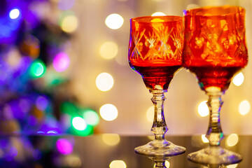 two beautiful red crystal glasses stand on a black glass table against the background of the Christmas tree and curtains of garlands with blurry background, used as a background or texture, soft focus