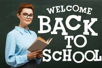 teacher in eyeglasses holding book near chalkboard with welcome back to school lettering in classroom