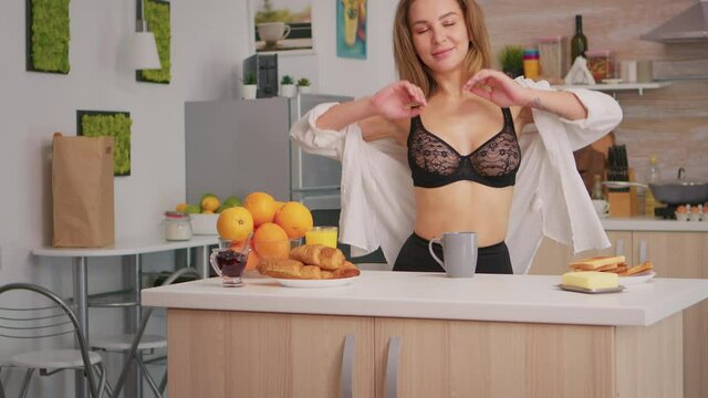 Blode beautiful woman wearing black lingeriesitting in the kitchen during breakfast. Young seductive attractive woman with tattoos in sexy underwear holding cup of coffee enjoying the morning smiling.