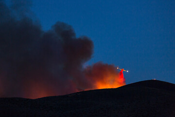 Firefighting plane soars from behind a hill dropping flame retardant. Last run of the night after sunset backlit smoke and fire - obrazy, fototapety, plakaty