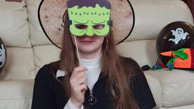 A smiling girl having fun with a paper mask, making funny and scary faces, get in ready for Halloween at home, celebrate Halloween at home with festive decorations
