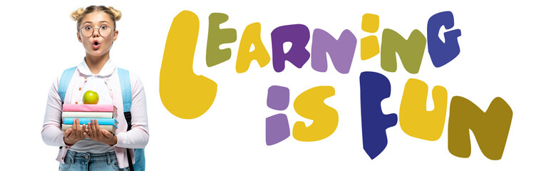 Panoramic shot of shocked girl with backpack holding books and apple near learning is fun lettering on white