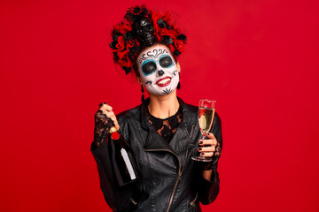 Girl with sugar skull makeup with a wreath of flowers on her head and skull, wearth lace gloves and leather jacket, Celebrates Halloween with a glass and a bottle of champagne in hands isolated on red