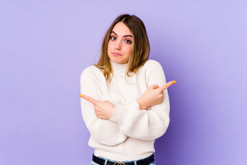 Young caucasian woman isolated on purple background points sideways, is trying to choose between two options.