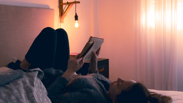 Momento de relajarse disfrutar del hogar y leer un buen libro en una c&oacute;moda c&aacute;lida y confortable habitaci&oacute;n.  Mujer adulta recostada en la cama leyendo un libro con sus pies posando sobre la pared