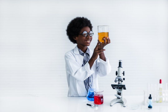 Happy African black boy in white gown student is learning and test science chemical with colorful liquid in beaker in laboratory room at to school. Education and science research concept.