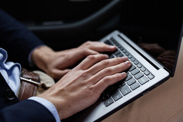 Close-up photo of male hands with laptop. Businessman in suit sitting in car, texting message, searching web, browsing information using laptop keyboard. Online education, remote working.