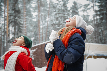 A warmly dressed brother and sister catch falling snowflakes with their tongue in winter in a snow park, during a family walk.