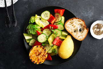Young boiled potatoes, salad of leafy greens, cucumber, capsicum, micro greens, oil, with lemon on a dark background in a black plate with a slice of baguette. Top view.