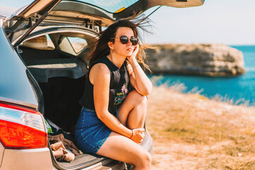 A young woman in sunglasses sits on the trunk of a car and looks cool, the sea coast in the background