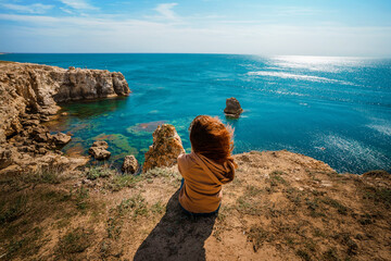 Fototapeta na wymiar Back view tourist woman enjoy seascape. Stylish girl sit on sea cliff and look at the ocean. 