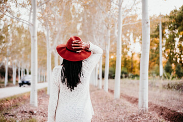 portrait of mid adult hispanic woman wearing a hat at sunset during golden hour, autumn season, beautiful path of trees background. back view