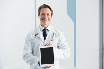 Doctor in white coat looking at camera and showing digital tablet with blank screen in hospital