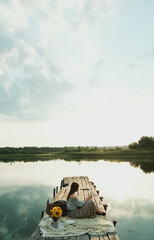 Fototapeta premium View of a Dreaming Girl Sitting on a Pier on a Lake and Dreaming. Sunrise on the Lake. Hot Tea, Teapot and Sweets Happiness or Inspiration Concept, Enjoy Life. Photo with Film and Grain Effect