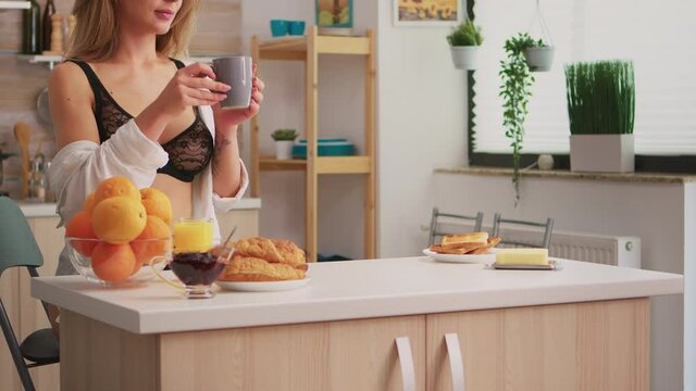 Attractive woman in black ligerie relaxing with coffee during breakfast sitting in kitchen and smiling. Young seductive blode lady with tattoos wearing sexy underwear refreshing in sunday morning