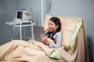 Little happy girl with an ice cream and a digital tablet in a hospital bed.