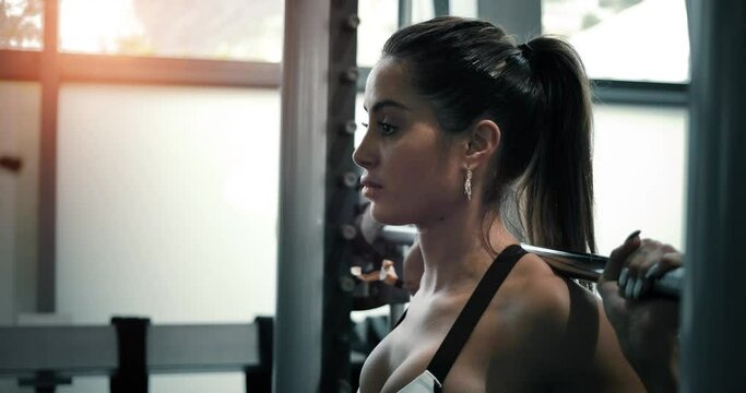 Woman is doing squats with a barbell in a fitness club. She is attractive and beautiful with long brown hair and she is focused on the exercice, with the sunlight in the background 4K