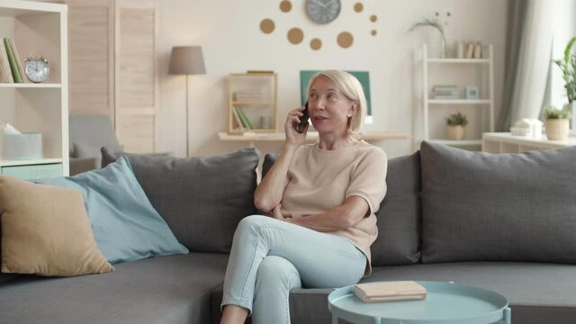 Medium pan shot of good-looking mature woman sitting on couch in living room chatting with kids on phone in middle of day