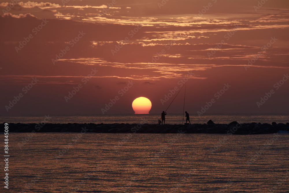 Wall mural The sun appears among the fishermen on the breakwater