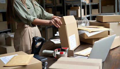 Female small business owner, entrepreneur, shipment delivery dropshipping service worker packing ecommerce order package in shipping cardboard box standing at table in warehouse. Close up view - obrazy, fototapety, plakaty