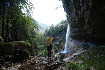 Young woman standing in front of waterfall with her hands outstretched. Caucasian female tourist in forest with her arms wide open