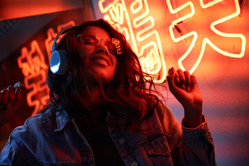 Happy cool fashion funky African young black hipster lady wears trendy glasses headphones listening dance pop dj music standing in neon night club light at style party. Translation: Chicken laugh