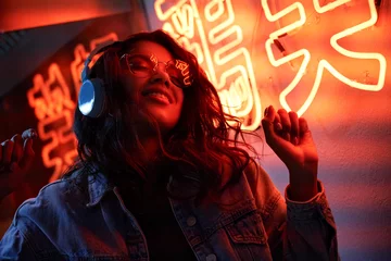 Gordijnen Happy cool fashion funky African young black hipster lady wears trendy glasses headphones listening dance pop dj music standing in neon night club light at style party. Translation: Chicken laugh © insta_photos