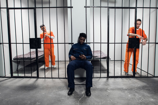 A strong black prison guard guards the cells with prisoners in the prison corridor.