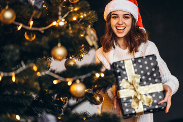 Pretty woman in warm sweater, socks and christmas hat, decorating new year tree and holding gif box