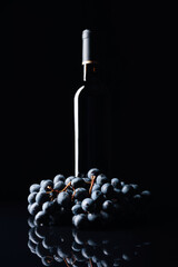 Bottle of red wine and a bunch of grapes on a black reflective background.