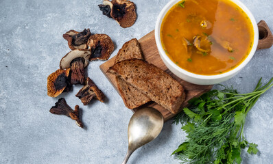 Mushroom soup with porcini mushrooms in a white round plate on a grey background. Nearby lies dark bread and dried porcini mushrooms. slow cooking