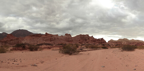 The arid desert in a hot day. Panorama view of the red sand, desert shrubs, sandstone and rocky formation in the background under a cloudy sky.