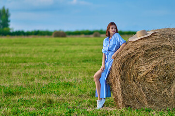 Naklejka na ściany i meble A girl in a blue dress near a haystack on a green field with a blue sky