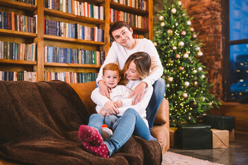 Christmas family portrait. Young Caucasian family in identical clothes having fun on sofa in living room with large library of books and Christmas tree in evening. Happy New Year and Merry Christmas