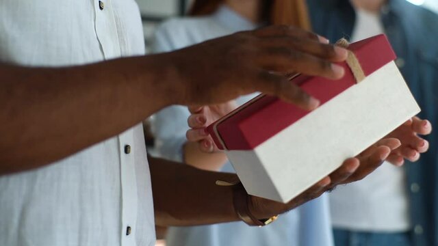 Young woman giving gift box to colleague, close-up of hands. Cheerful birthday man opening the box with gift and surprised. African-American male showing joy. Shooting in slow motion.