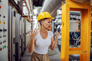 Angry hardworking female supervisor in working suit with protective helmet on head yelling over walke talkie on her employee. Mistake can't happen.