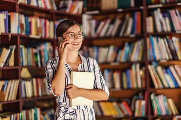 Young smiling attractive college girl with eyeglasses and in dress standing in library, holding book and having phone call.