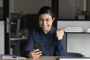 Excited millennial indian woman sit at desk look at smartphone feel euphoric win lottery online....