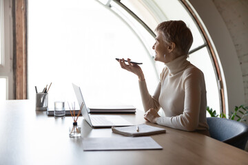 Smiling young Caucasian businesswoman sit at desk at workplace record audio message use virtual...